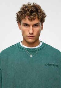 Groen, oversized sweatshirt met textuur, geborduurde details en een ronde hals, gelaagd over een wit shirt, gecombineerd met gelaagde zilveren kettingen.