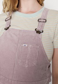 Lila kordflodsoveraller med en randig mint- och gräddvit skjorta under, med metallhardware och en liten "Lee"-logotyp på fickan.