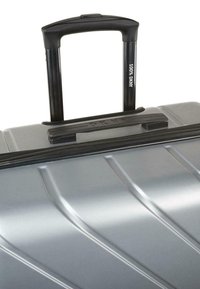 Maleta rígida plateada con una superficie texturizada, que cuenta con un asa retráctil negra y un cierre. Marcada con "DKNY" en el asa.