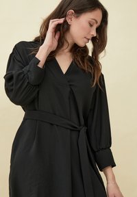 Zwarte wikkeljurk met lange, opgeblazen mouwen, een v-hals en een strikceintuur in de taille. Gemaakt van een gladde, lichtgewicht stof met een matte afwerking.
