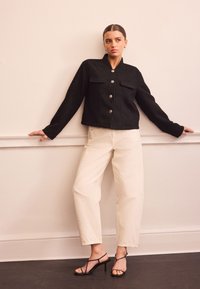 Giacca nera corta con bottoni coordinati e due tasche frontali, abbinata a pantaloni a gamba larga color crema e tacchi neri con cinturini.