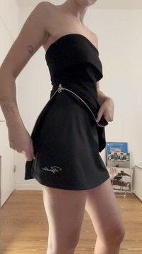 Fekete, pánt nélküli ruha, testhez simuló felsőrésszel és bővülő szoknyával, ezüst oldalsó cipzárral és fehér hímzett logóval az alsó szegélynél.
