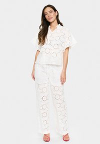 Conjunto de dos piezas blanco que incluye una blusa de manga corta con capucha y pantalones de pierna ancha, ambos con patrones de ojales y bordes festoneados.