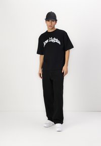 Fekete, oversize t-shirt, amelyen fehér felirattal olvasható a "low lights" szöveg, fekete nadrággal és fehér tornacipővel párosítva. Minimalista dizájn, semleges háttéren.
