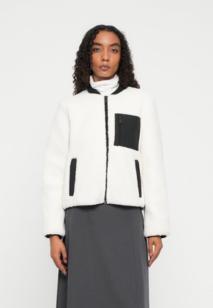 Eine Frau trägt eine weiße Fleece-Zip-Jacke mit schwarzen Taschen und Kragen, kombiniert mit einem dunkelgrauen Rock vor einem schlichten weißen Hintergrund.
