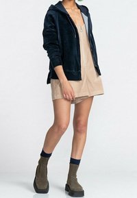 Navy corduroy zip-up hoodie with a front pocket, paired with a beige short dress featuring a central zipper. Olive green ankle boots.