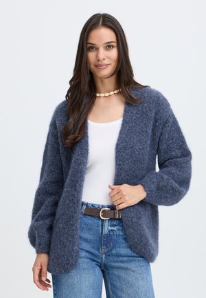 Frau mit langen braunen Haaren trägt eine blaue, flauschige Strickjacke, ein weißes Oberteil, blaue Jeans mit Gürtel und eine Halskette, steht vor einem schlichten Hintergrund.