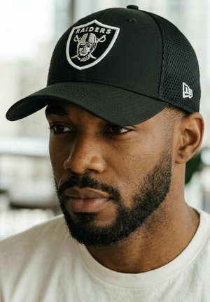 Hombre negro con barba, que lleva una gorra de béisbol negra de los Raiders y una camiseta blanca, mirando ligeramente hacia un lado con luz suave en interiores.
