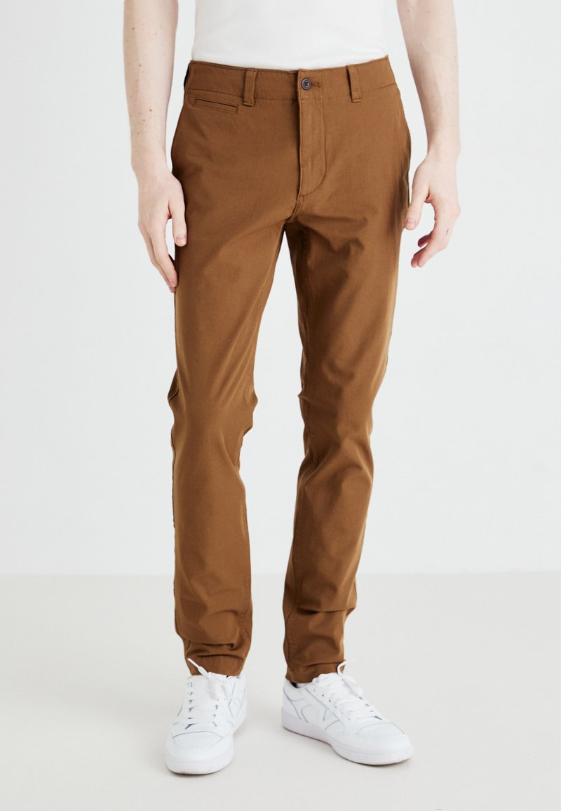 Brune bomullsk chinos med smal passform, med sidelommer, en enkelt knappelukking og en glatt tekstur, kombinert med hvite sneakers.