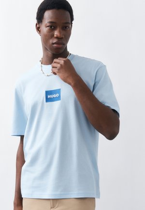 Νέος άνδρας που φοράει ανοιχτό μπλε μπλουζάκι HUGO και κολιέ με χάντρες, κρατώντας το κολιέ με το δεξί χέρι σε μονόχρωμο φόντο.