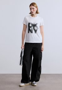 Άσπρο βαμβακερό T-shirt με μεγάλο μαύρο παγιετένιο τύπωμα "DKNY", συνδυασμένο με μαύρο cargo παντελόνι και ασημένια αθλητικά, κρατώντας μια ασημένια τσάντα tote.