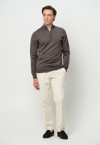 Brauner Pullover mit langen Ärmeln und halber Zip-Kragen, kombiniert mit hellen Hosen und dunklen Slippern; glatte Textur.