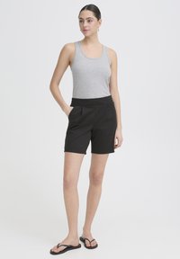 Žena stojaca, oblečená v sivom tielku bez rukávov, čiernych krátkych nohaviciach s vysokým pásom a vreckami a čiernych žabkách na bielom neutrálnom pozadí.