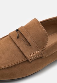 Brown suede slip-on shoe featuring a flap with a cutout design, decorative stitching along the edges, and a smooth inner lining.