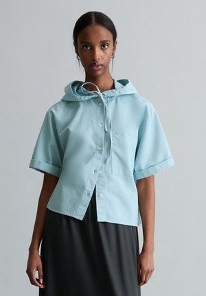 Woman with dark skin wearing a light blue short-sleeve hooded button-up shirt over a black dress, standing against a plain background.