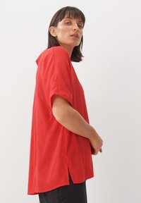 Rotes, lockeres kurzärmliges Shirt mit rundem Ausschnitt. Hergestellt aus leichtem Stoff, mit Seitenschlitzen am Saum für zusätzlichen Bewegungsfreiheit.