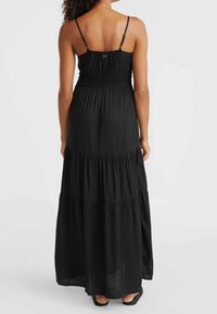 Kvinna som bär en lång svart ärmlös klänning med justerbara remmar, står barfota mot en enkel ljus bakgrund.