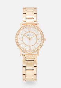 Auksinės spalvos Guess laikrodis su apvaliu korpusu, baltu ciferblatu, kristaliniais akcentais ir sujungtu apyrankės dirželiu. Pasižymi drąsiais arabų skaičiais ir valandų žymekliais.