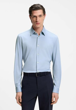 Hombre que lleva una camisa azul claro abotonada y metida dentro de pantalones oscuros con un cinturón negro, de pie contra un fondo blanco liso.