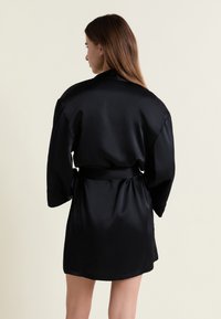 Femme aux longs cheveux bruns vue de dos, portant un peignoir en satin noir à larges manches et ceinture à la taille, sur un fond clair uni.