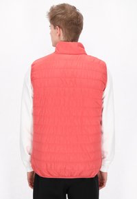 Gilet imbottito in vivace corallo, con colletto alto e texture trapuntata. Indossato sopra una camicia a maniche lunghe bianca, con orlo elasticizzato.