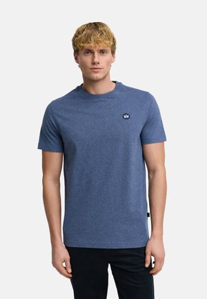 Blonder Mann trägt ein schlichtes blaues Kurzarm-T-Shirt mit einem kleinen runden Logo auf der linken Brust, steht vor einem einfarbigen hellen Hintergrund.