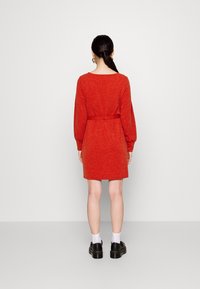 Vestido de punto rojo con mangas largas, cintura ajustada y longitud hasta mitad del muslo. Lleva zapatos negros y calcetines blancos. Textura suave.