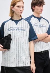 Νεαρή γυναίκα κρατώντας μαύρη τσάντα και νεαρός άνδρας με σταυρωμένα τα χέρια, φορούν λευκά και ναυτικά μπλουζάκια με κείμενο "Carlo Colucci".