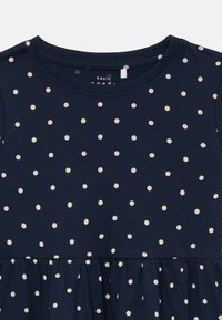 Abito blu navy con pois crema, caratterizzato da un colletto rotondo e gonna arricciata. Realizzato in un tessuto morbido e leggero.