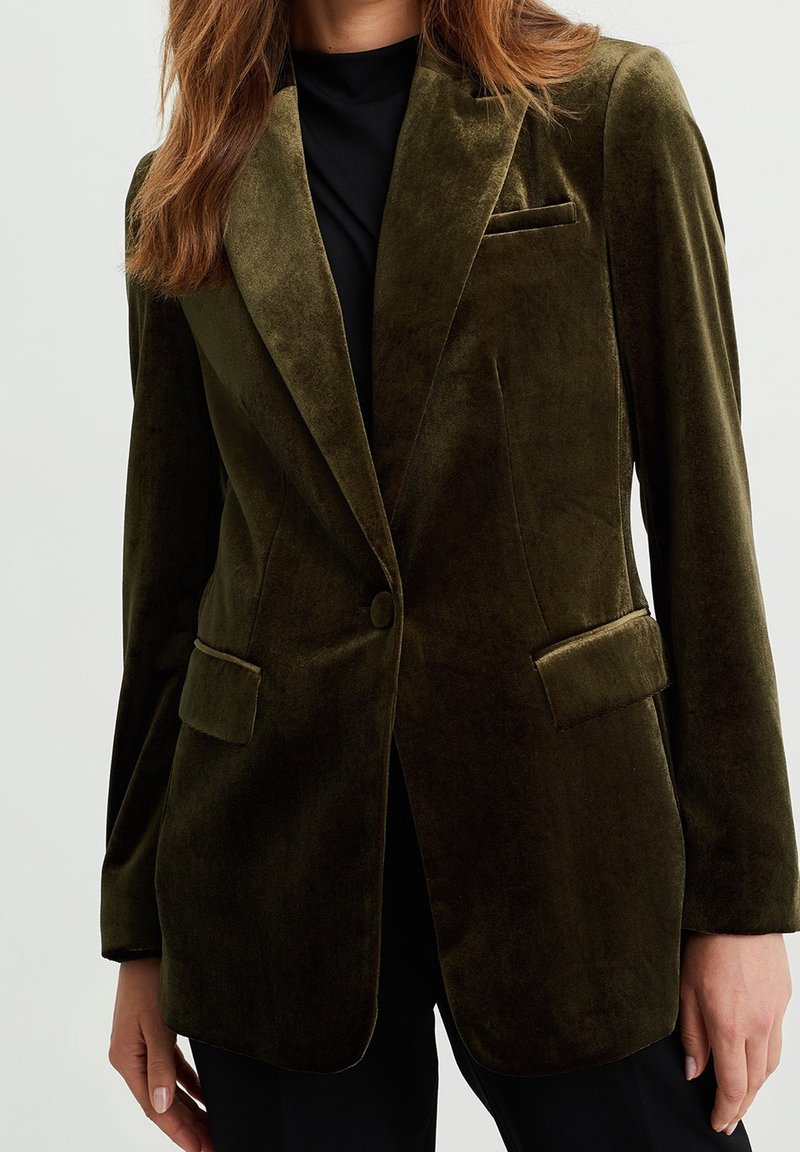 Femme portant un blazer en velours vert olive avec revers crantés, bouton unique, poches à rabat, et un haut noir en dessous, sur fond uni.