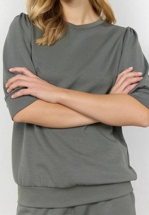 Personne portant un haut vert pâle à manches courtes avec des détails froncés aux épaules, les bras croisés sur la poitrine devant un fond uni.