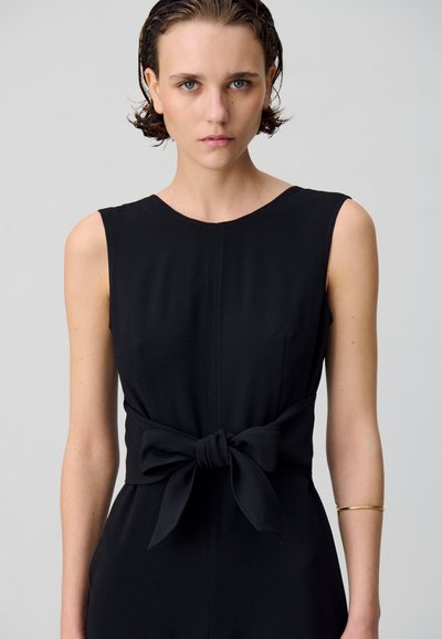 Robe noire sans manches avec un col rond, ornée d'un nœud noué à la taille. Tissu lisse avec des accents de couture subtils.