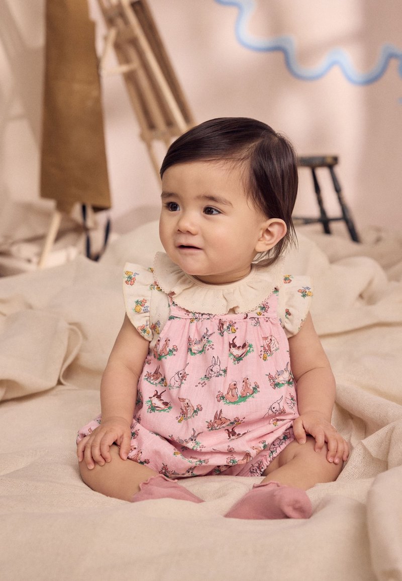 Salopetă roz cu imprimeu floral, cu guler crem cu volane, decorată cu imprimeuri de animale, asortată cu șosete roz, pe un fundal de material deschis la culoare.