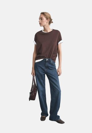 Brown t-shirt with white cuff accents, paired with blue straight-leg jeans. Model holds a dark handbag, wearing dark slip-on shoes.