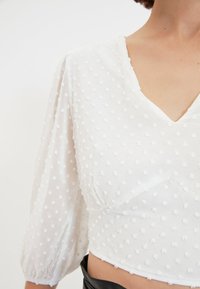 Blusa bianca con maniche a sbuffo e scollatura a V, caratterizzata da un motivo a pois testurizzati. Il tessuto è leggero e trasparente.