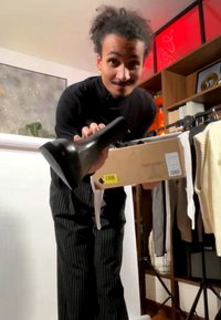 Homme en chemise noire et pantalon à rayures tient une botte noire et une boîte à chaussures dans une pièce avec des vêtements et des étagères à livres.