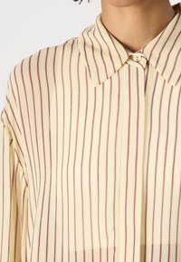 Žltá pruhovaná blúzka s tenkými bordovými vertikálnymi pruhmi, s vystúpeným golierom a zapínaním na gombíky pri výstrihu, hladká textúra látky.