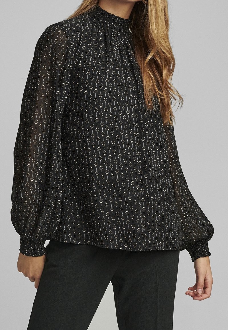 Blusa nera con collo alto arricciato, maniche a sbuffo lunghe e un delicato motivo di cerchi dorati su tutta la stoffa.