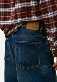 Jeans in denim di colore blu scuro con design classico a cinque tasche. Presenta una patch in pelle color beige con l'etichetta "TOMMY JEANS" e sottili accenti rossi, bianchi e blu.