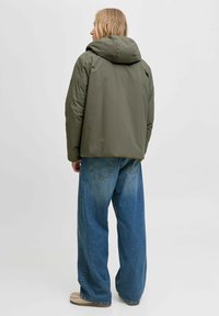 Olivgrüne Pufferjacke mit Kapuze, glatte Textur, leicht verkürzter Schnitt, kombiniert mit lockeren blauen Jeans, hell ausgewaschen, und lässigem Schuhwerk.