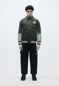 Sötétzöld dzseki szürke ujjakkal, gombos elülső rész, csíkos mandzsettákkal; fekete cargo nadrággal és fekete sportcipővel párosítva.