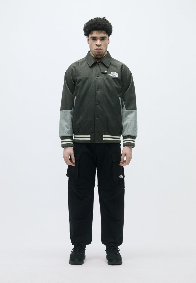 Sötétzöld dzseki szürke ujjakkal, gombos elülső rész, csíkos mandzsettákkal; fekete cargo nadrággal és fekete sportcipővel párosítva.