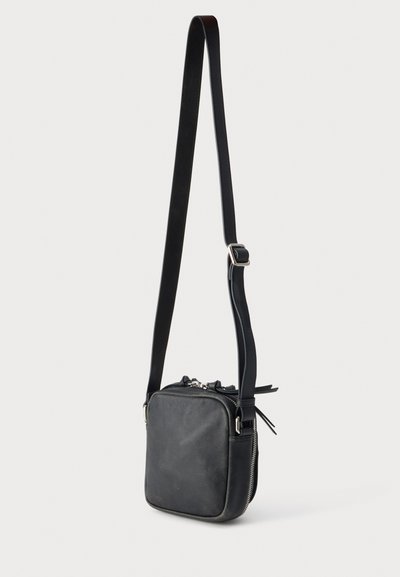 Μικρό μαύρο δερμάτινο τσαντάκι crossbody με ρυθμιζόμενο λουράκι και ασημένια φερμουάρ σε απλό φωτεινό φόντο.