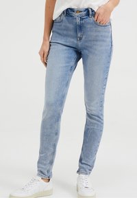 Vysoké modré džíny z denimu s přiléhavým střihem, pěti kapsami a zapínáním na knoflík, v kombinaci s bílými teniskami.