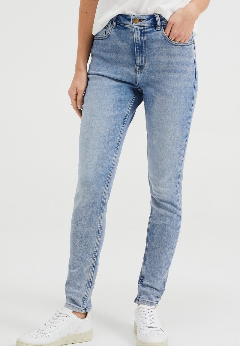 Vysoké modré džíny z denimu s přiléhavým střihem, pěti kapsami a zapínáním na knoflík, v kombinaci s bílými teniskami.