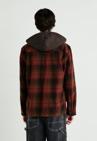 Camicia a quadri nei colori rosso, nero e marrone; presenta un cappuccio marrone e polsini con bottoni, mostrando un tessuto testurizzato con motivi variabili.