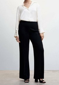 Chemise blanche à manches longues avec col, associée à un pantalon large noir taille haute et des escarpins ouverts noirs sur un fond neutre.