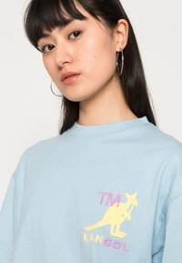 T-shirt de algodão azul claro com um gráfico de canguru e o texto "TM KANGOL" em bordado rosa e amarelo na área do peito esquerdo.