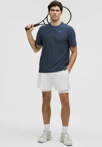 Conjunto atlético para hombres que incluye una camiseta de manga corta azul oscuro y pantalones cortos blancos. La camiseta tiene una textura suave y un logo en el pecho.