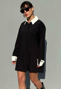 Zwarte oversized shirtjurk met een witte kraag en manchetten, voorzien van knoopsluiting en decoratieve knopen op de mouwen. Glad stof.